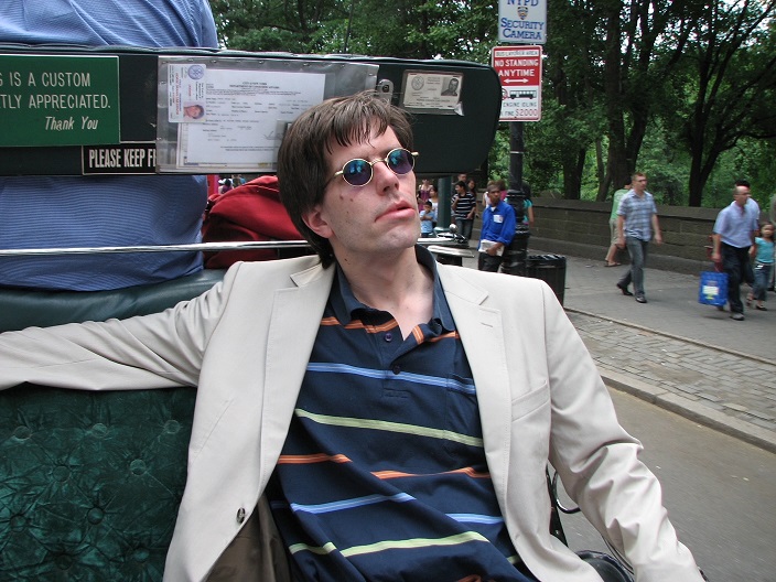 Lars Bruhn, ein schlanker Mann mit kurzen schwarzen Haaren und Sonnenbrille, sitzt auf einer Parkbank bei sonnigem Wetter und blickt in die Ferne. Er trägt ein blaues Hemd und eine beige Anzugsjacke. [Foto: private Aufnahme]